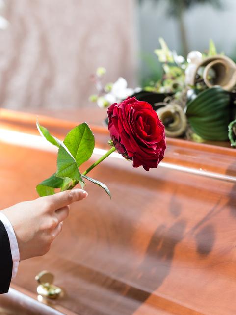 Frau in schwarzem Anzug hält eine rote Rose über einen Holzsarg, der mit Blumen dekoriert ist.