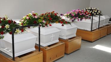 Reihe weißer Särge mit Blumenarrangements auf Holzständern in einem hellen Raum.