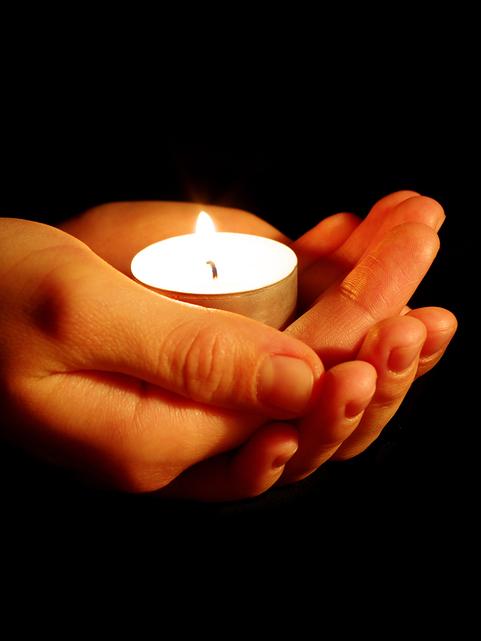 Hände halten eine flackernde Teelichtkerze vor einem dunklen Hintergrund.