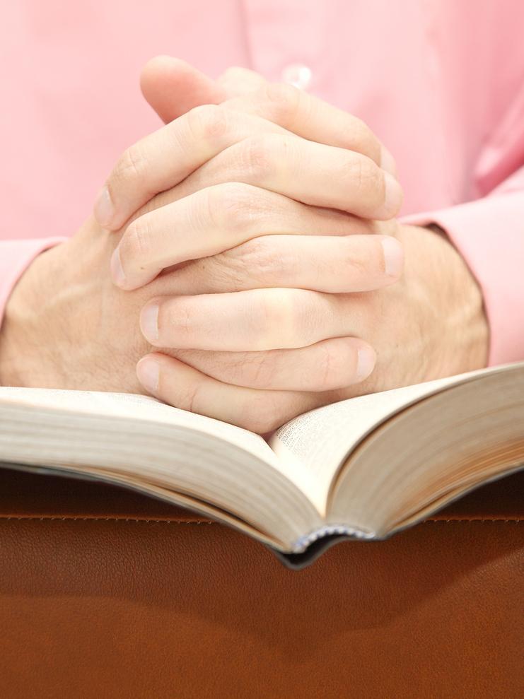 Hände eines Mannes sind gefaltet über einem offenen Buch, auf einem braunen Ledersofa. Der Mann trägt ein rosa Hemd.