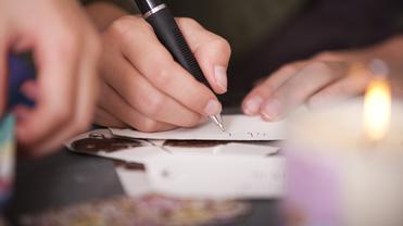 Nahaufnahme einer Hand, die mit einem Kugelschreiber auf einem Zettel schreibt, im Hintergrund eine brennende Kerze.